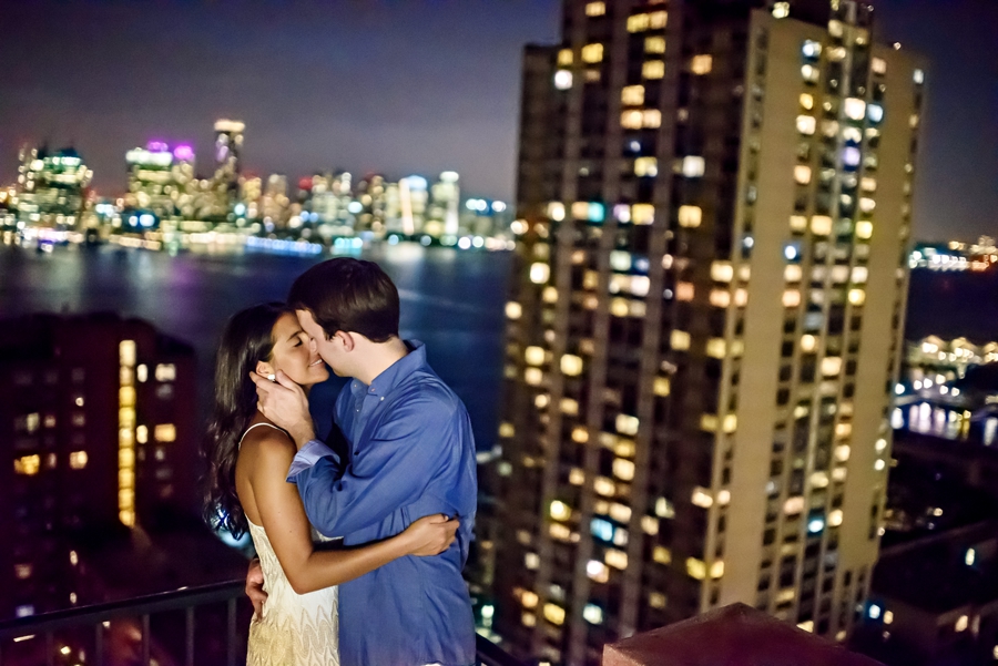 An engaged couple kiss on the roof of thier NYC apartment building with the hudson river in the background during their NYC engagement session.